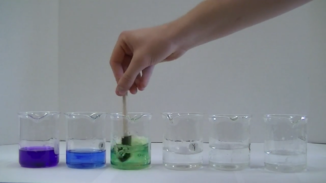 Demonstrators hand adding solution to series of beakers with a variety of solution colors